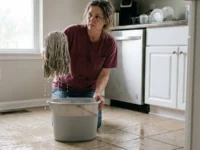 A woman holding a dripping wet string mop over a bucket of gray water on a wet streaky kitchen tile floor