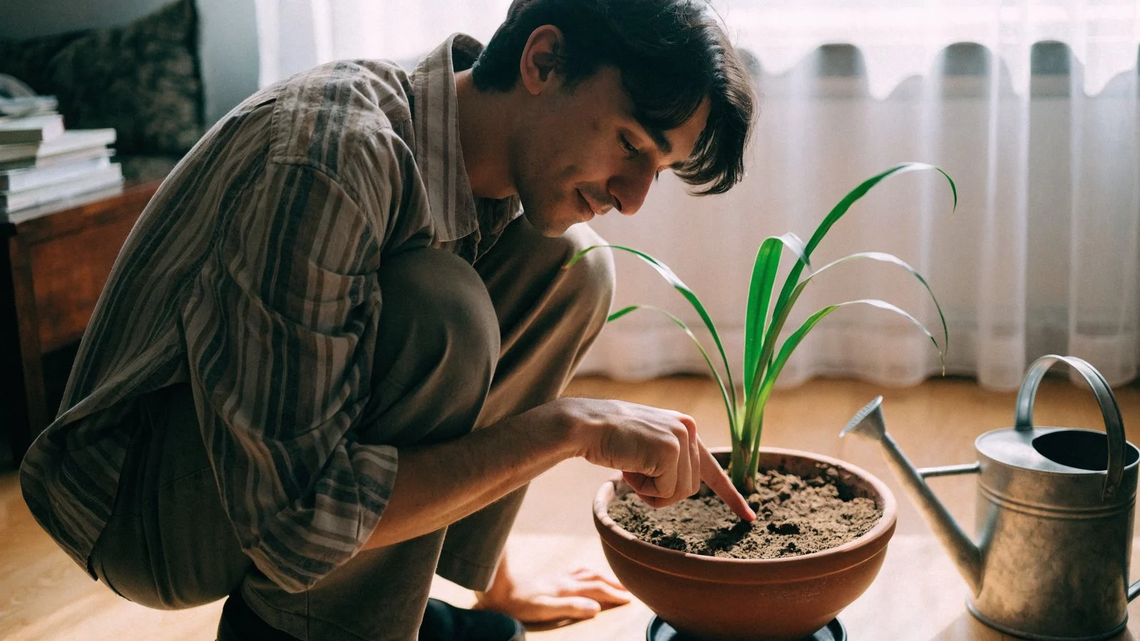Man crouching beside a potted plant pressing a finger into dry soil to test moisture, with a watering can set aside.